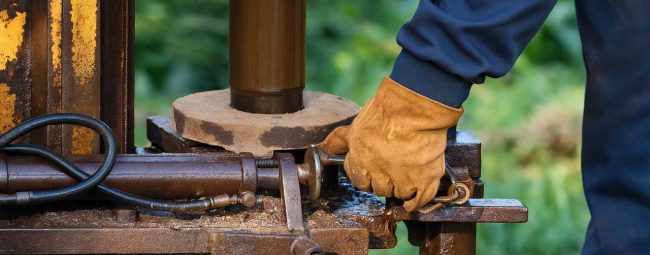 A person wearing a blue long-sleeve shirt and yellow gloves is adjusting a mechanical component on a rusted metal structure, set against a green outdoor background.