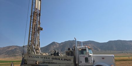 A white Evans Well Drilling truck with its large drilling mast extended, positioned in a dry field with mountains in the background under a clear blue sky.