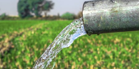 A close-up of water gushing out of a pipe, highlighting its flow into a lush green field with trees in the distance.