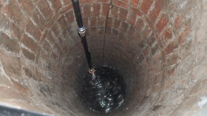 The interior of a brick-lined well shaft, showing a black pipe extending downward into water at the bottom.