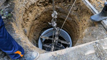A deep well is being installed, with a cylindrical component suspended by ropes and chains, lowered into an excavated hole.