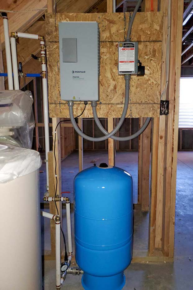 A blue water storage tank is connected to pipes and valves, part of a residential water pumping system installed in an unfinished room.