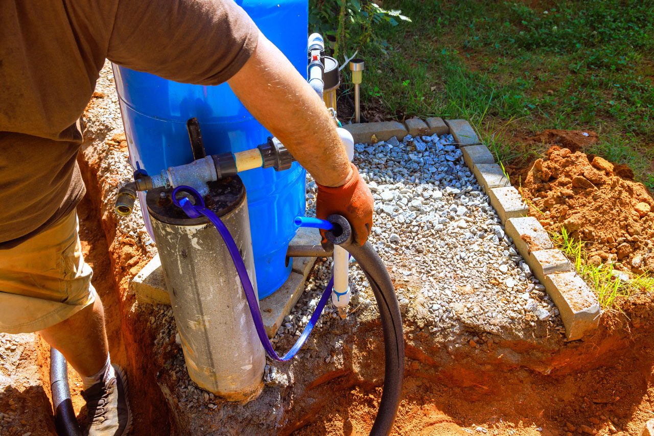 A worker wearing gloves lowers a submersible water pump into a well casing next to a large blue pressure tank during an installation.