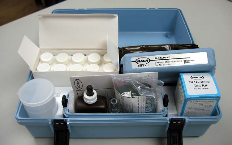 A blue testing kit containing various reagents, bottles, and instructions for measuring water alkalinity and hardness is displayed on a table.